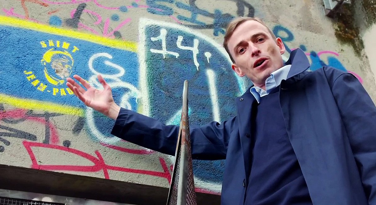 Man facing camera and pointing to a wall with Graffiti on it 