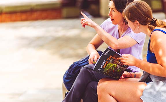 Two women sit in one of Newcastle Universities quads, reading through the Summer Open Day booklet.