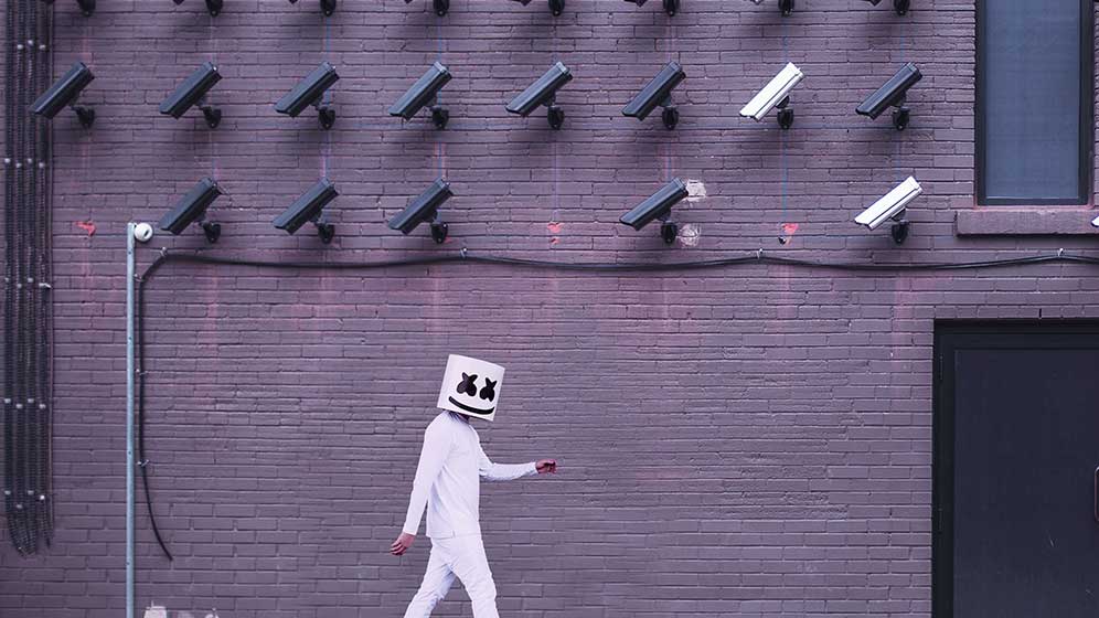 Person in white hazmat suit walking past a wall of security cameras.