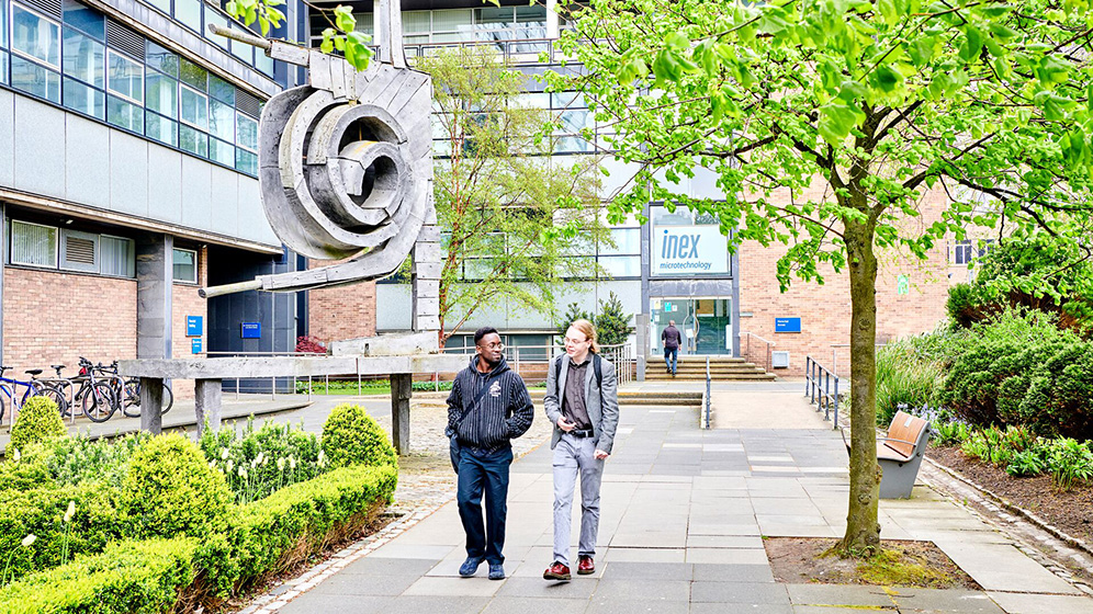 Two students walking together past the statues outside the Herschel Building.
