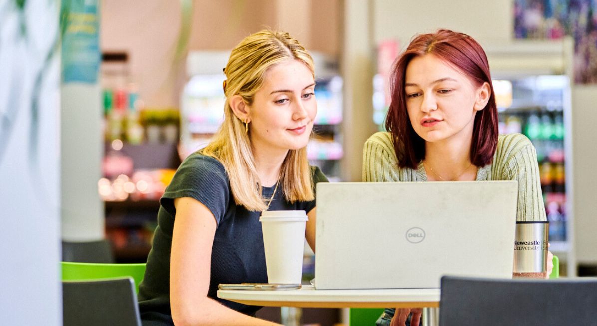 Two students at a NUBS café table study a laptop screen together.